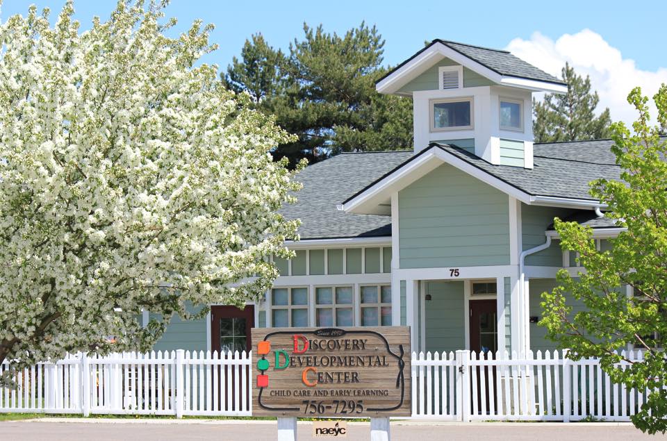 Image of a one-level school house with bell tower, surrounded by trees. School is painted sea-foam green with white trim and white picket fence. A wooden sign out front reads "Discovery Developmental Center"