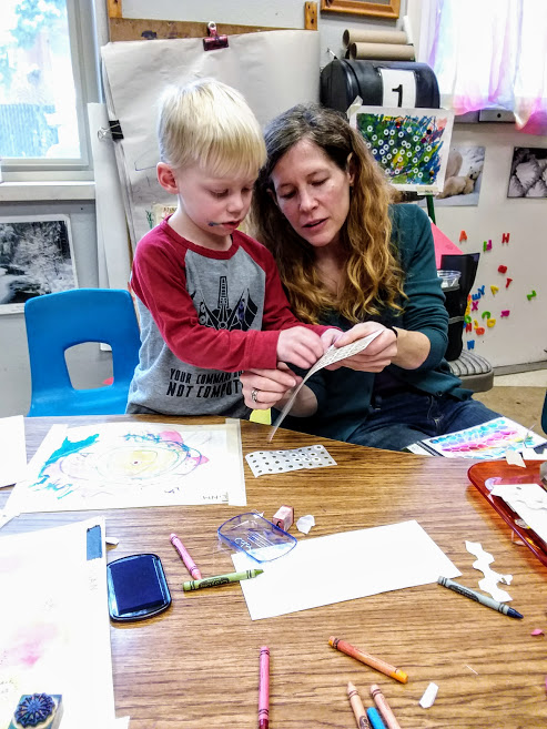 A woman helps a young boy peel stickers from a sticker sheet. They are sitting at a low table that is covered in crayons, paper, stamp pads, and stamps.