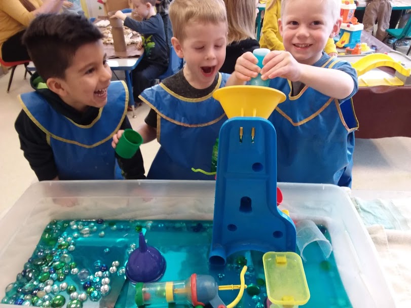 Three smiling boys wearing paint smocks stand over a shallow tub full of blue dyed water and water toys. One boy squeezes a bottle of blue water into a funnel while the other two watch.