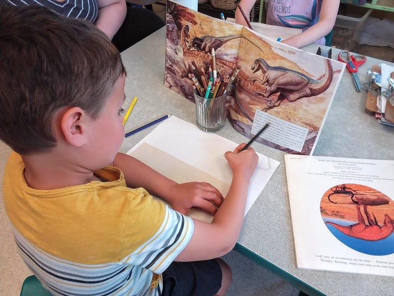 A young boy holds a pencil and works to draw a picture, referencing two dinosaur picture books on the table. 